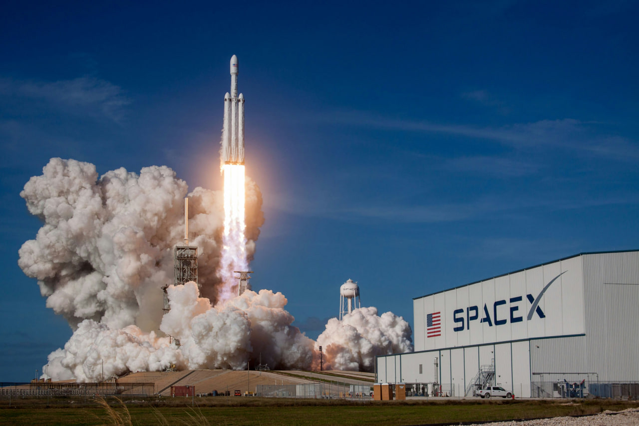 Запуск ракеты SpaceX Falcon Heavy с площадки, облака дыма и здание SpaceX на переднем плане